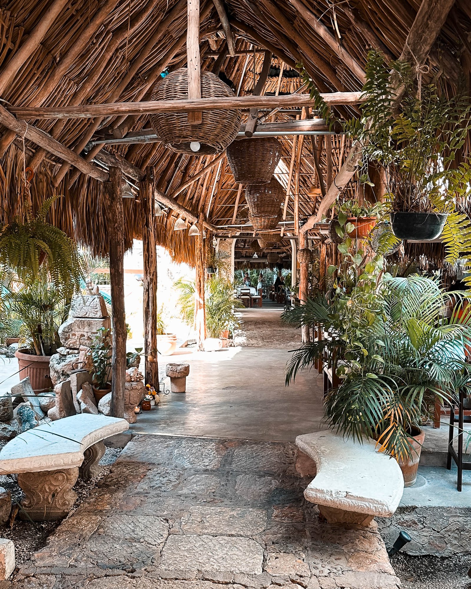 Eingangsbereich eines Restaurants der mit einem Naturdach bedeckt mit Palmenblättern beschützt wird. Entlang des Weges stehen Sitzgelegenheiten