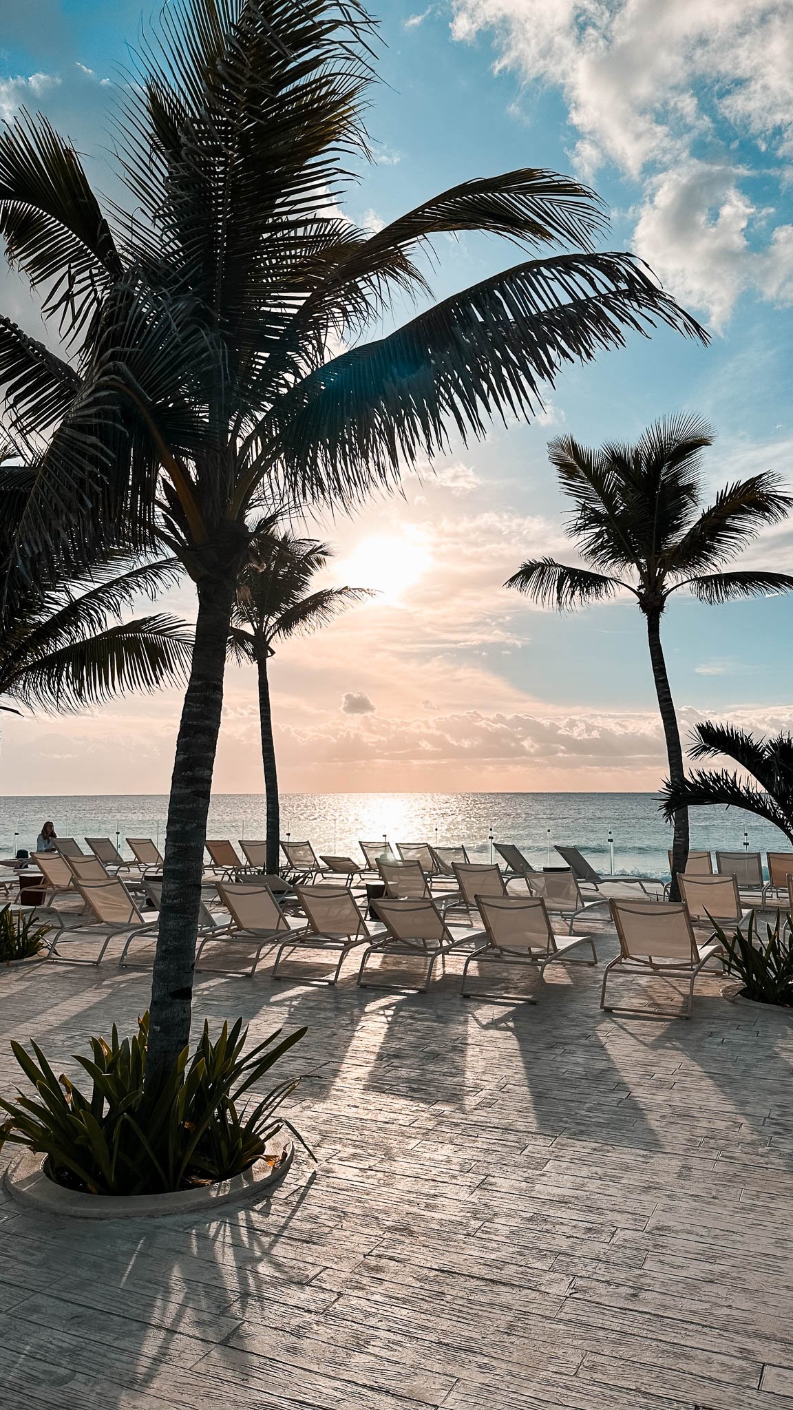 Strandbereich mit liegen und Palmen dahinter geht die Sonne gen Meer unter