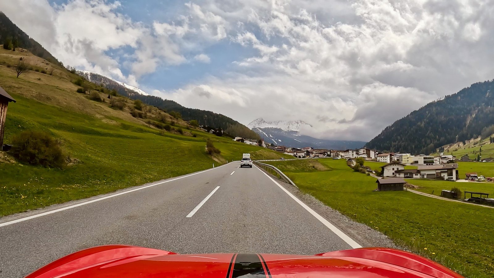 Perspektive des Fahrers zeigt den Blick auf ein Dorf mit einem schneebedeckten Berg im Hintergrund am Reschenpass